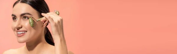 Щаслива молода жінка робить масаж обличчя з нефритним роликом ізольовано на рожевому, банер — стокове фото