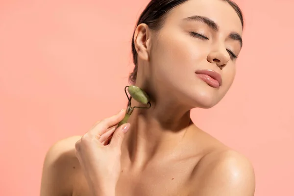 Brunette young woman doing neck massage with jade roller isolated on pink — Stock Photo