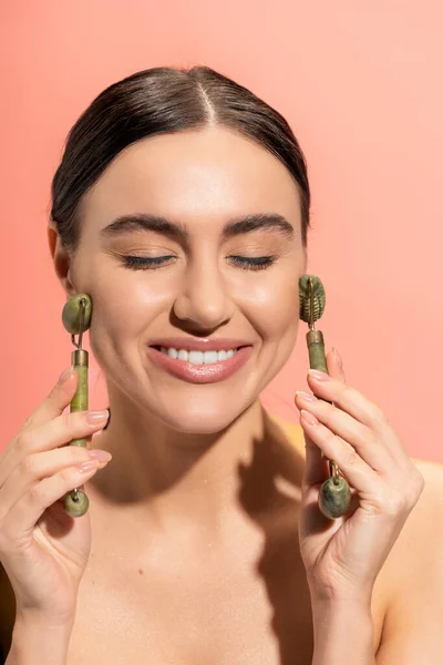Pleased woman doing face massage with jade rollers isolated on pink — Stock Photo