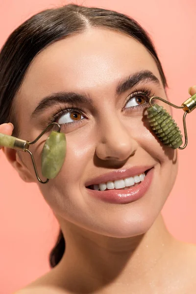 Close up view of cheerful young woman doing face massage with jade rollers isolated on pink — Stock Photo