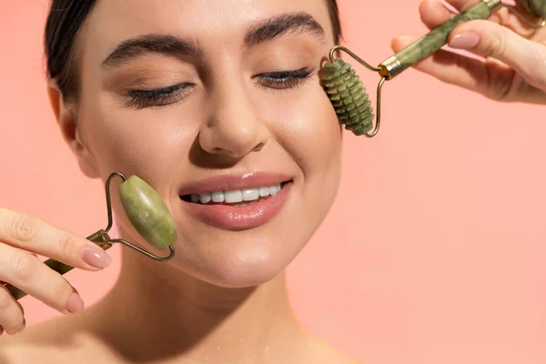 Close-up de jovem mulher positiva fazendo massagem facial com rolos de jade isolados em rosa — Fotografia de Stock