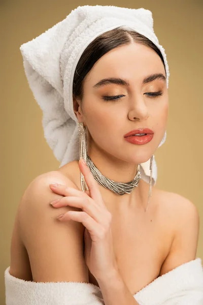 Elegant young woman with makeup and towel on head touching bare shoulder on beige background — Stock Photo