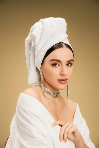 Young woman in jewelry with white towel on head looking at camera on beige background — Stock Photo