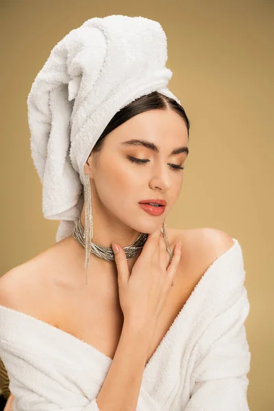 Charming woman in jewelry with white towel on head touching chin on beige background — Stock Photo