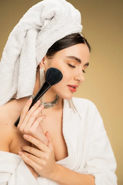 Mujer joven con toalla blanca en la cabeza aplicando polvo facial con cepillo de maquillaje sobre fondo beige - foto de stock