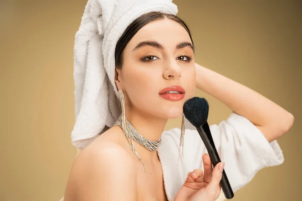 Pretty woman in white towel on head applying face powder with makeup brush on beige background — Fotografia de Stock