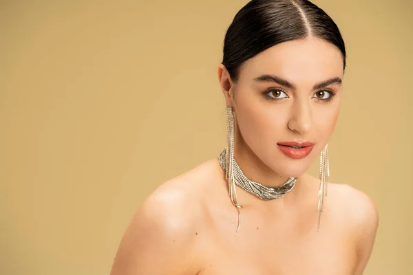 Brunette young woman in necklace and earrings looking at camera isolated on beige — Stock Photo