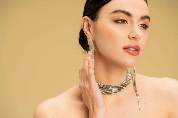 Brunette young woman in necklace adjusting earrings while looking away isolated on beige — Stock Photo