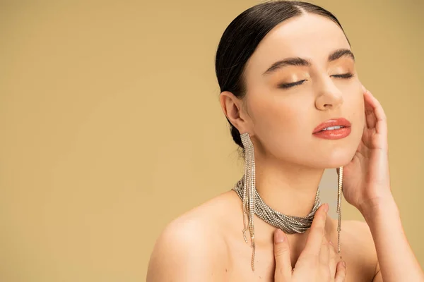 Brunette young woman in necklace and earrings posing with closed eyes isolated on beige — Stock Photo