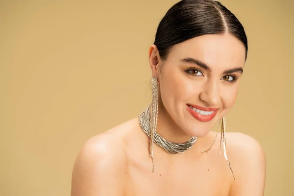 Joyful woman in necklace and earrings looking at camera isolated on beige — Stock Photo