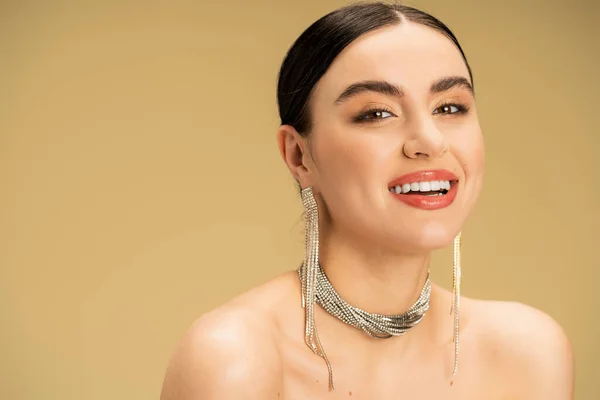 Elegant woman in necklace and earrings looking at camera while smiling isolated on beige — Fotografia de Stock