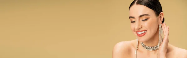 Morena joven en collar y pendientes sonriendo aislado en beige, pancarta - foto de stock