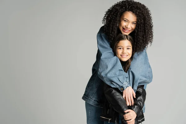 Happy curly mother hugging stylish kid in leather jacket isolated on grey — стоковое фото