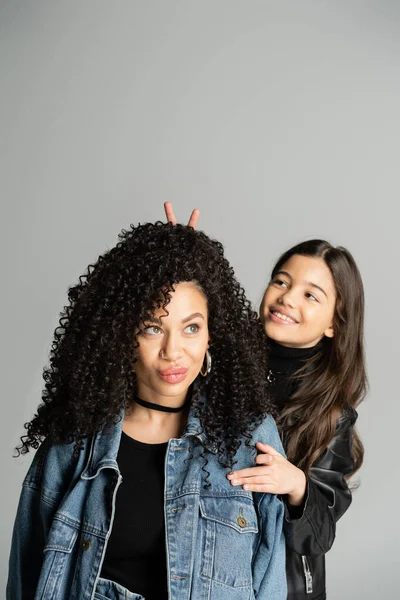 Smiling kid showing bunny ears with fingers near curly mom pouting lips isolated on grey — Stock Photo