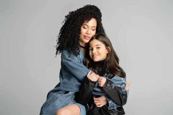 Curly woman in denim jacket hugging happy daughter isolated on grey — Stock Photo