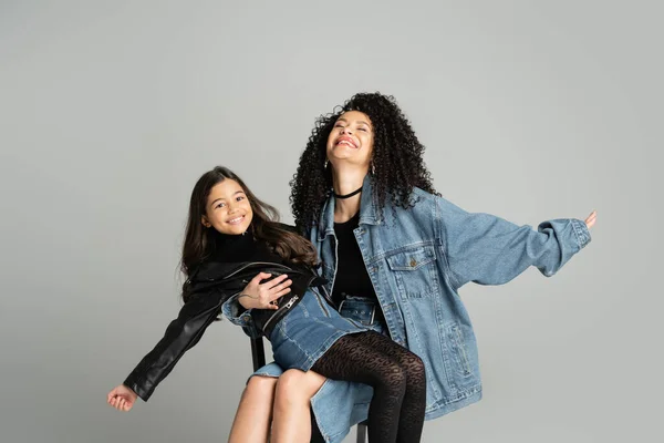 Mujer positiva en chaqueta vaquera sosteniendo hija alegre aislada en gris - foto de stock