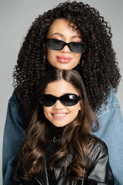 Portrait of curly woman in sunglasses posing near smiling daughter isolated on grey — стоковое фото