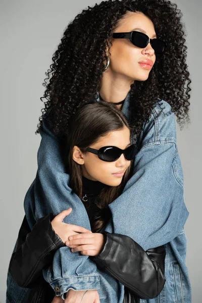 Fashionable woman in sunglasses hugging daughter isolated on grey - foto de stock