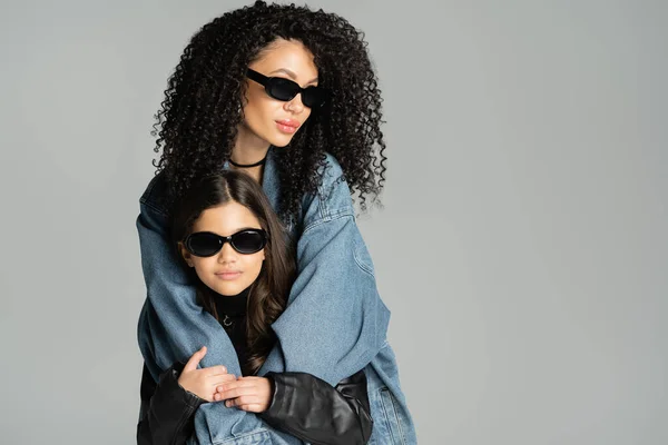 Trendy curly woman in sunglasses hugging preteen kid isolated on grey - foto de stock