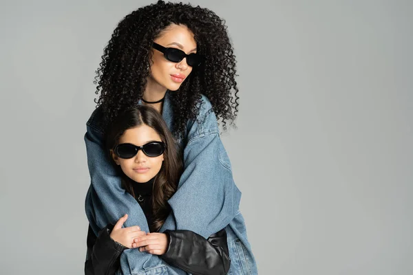 Trendy curly woman in sunglasses hugging preteen kid isolated on grey — стоковое фото