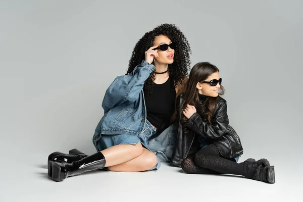 Trendy woman and happy daughter in sunglasses sitting on grey background - foto de stock