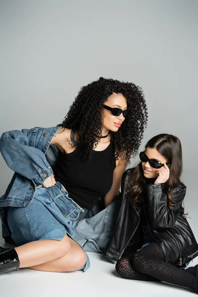 Stylish woman in denim jacket and sunglasses sitting near daughter on grey background - foto de stock
