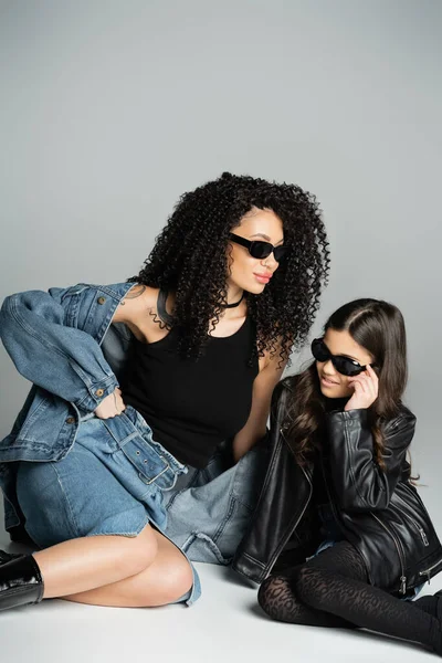 Stylish woman in denim jacket and sunglasses sitting near daughter on grey background — стоковое фото