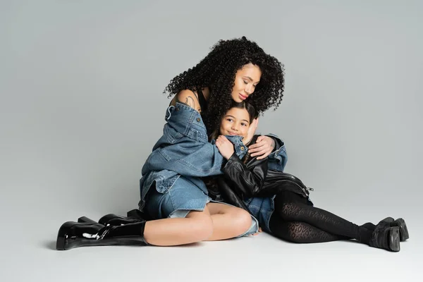 Tattooed woman in denim jacket hugging smiling child on grey background — стоковое фото