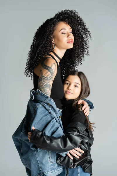 Tattooed young woman in denim jacket posing with child isolated on grey — стоковое фото