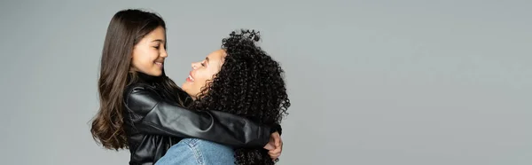 Side view of smiling kid in leather jacket hugging curly mom isolated on grey, banner — Photo de stock