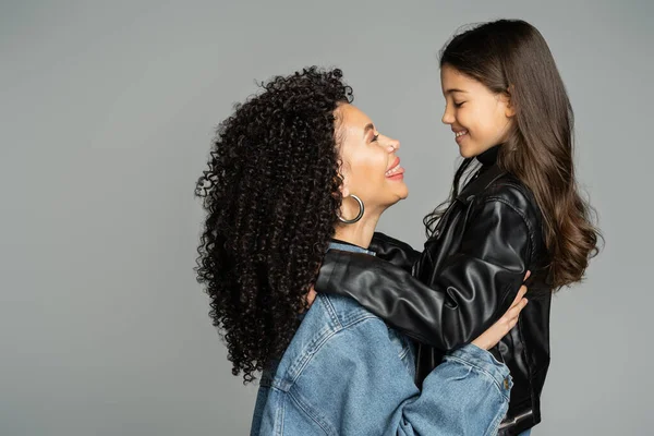 Vista lateral de mamá alegre en chaqueta de mezclilla que sobresale de la lengua mientras abraza a la hija aislada en gris - foto de stock