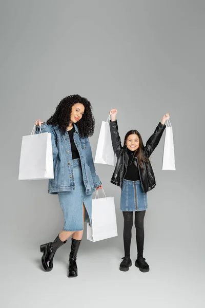 Full length of stylish mother and daughter holding shopping bags on grey background — Photo de stock
