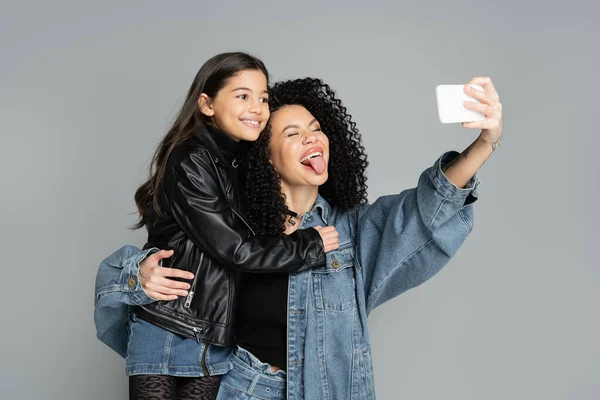 Stylish woman sticking out tongue while taking selfie with daughter isolated on grey — Stock Photo