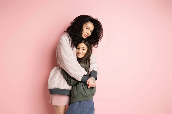 Donna riccia in bomber giacca abbracciando figlia su sfondo rosa — Foto stock
