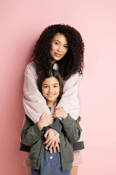 Curly woman hugging preteen girl on pink background - foto de stock