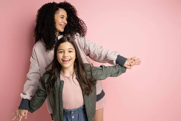 Positive curly woman holding hand of preteen daughter on pink background - foto de stock