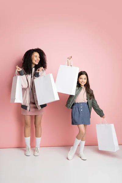 Cheerful mother and daughter holding shopping bags on pink background — Stockfoto