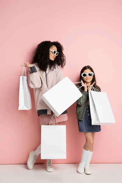 Stylish preteen girl holding purchases near mom in sunglasses on pink background — Stockfoto