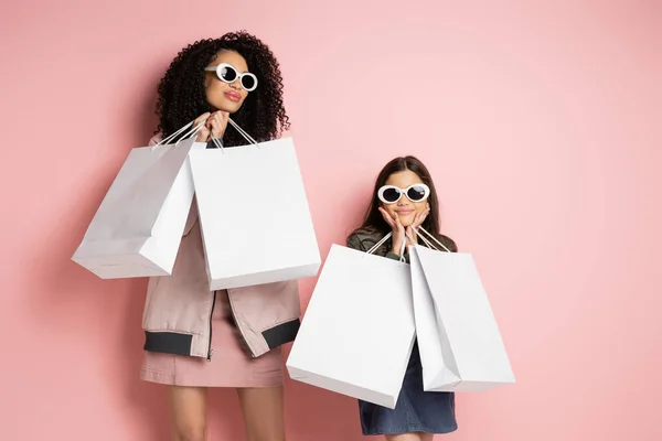 Fashionable mother and daughter in sunglasses holding shopping bags on pink background — Photo de stock
