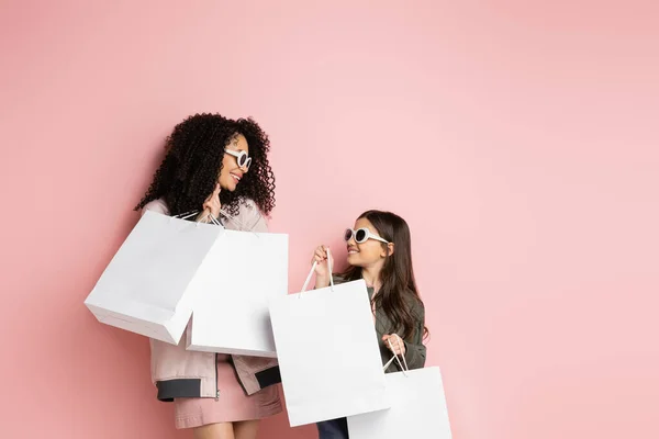 Positive and stylish woman holding shopping bags near daughter on pink background — Stockfoto