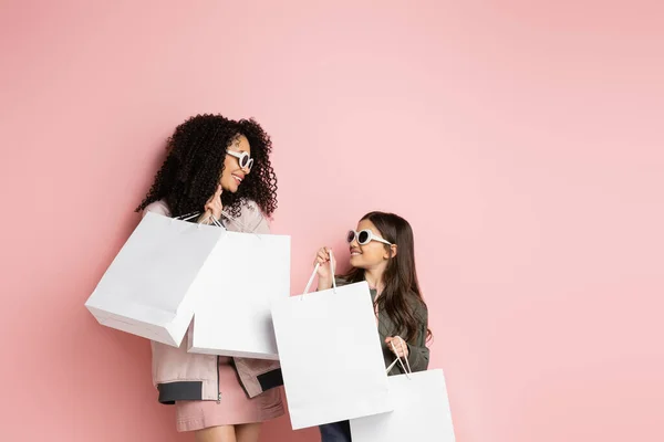 Mujer positiva y elegante sosteniendo bolsas de compras cerca de la hija sobre fondo rosa - foto de stock