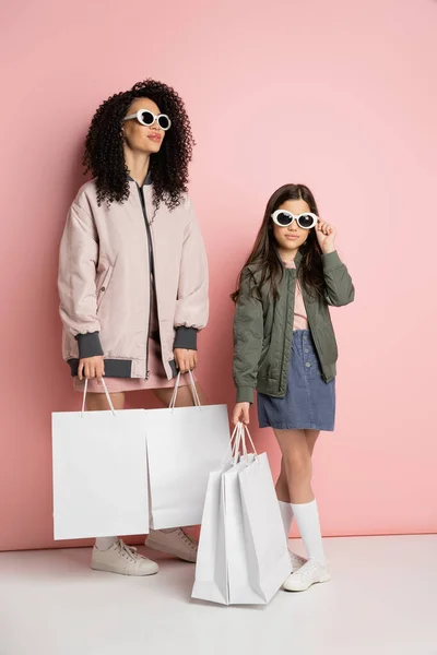 Fashionable mother and kid in sunglasses holding shopping bags on pink background — Photo de stock