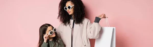 Trendy madre in occhiali da sole in possesso di shopping bag vicino figlia su sfondo rosa, banner — Foto stock