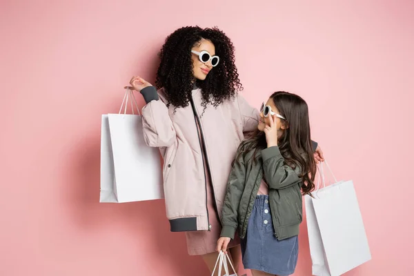 Donna alla moda e bambino in occhiali da sole che tengono borse della spesa su sfondo rosa — Foto stock