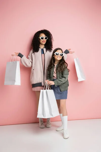 Full length of trendy mother and daughter in sunglasses holding purchases on pink background - foto de stock