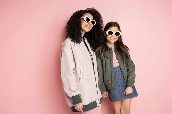 Positive Mutter und stilvolle Tochter mit Sonnenbrille halten Händchen auf rosa Hintergrund — Stockfoto