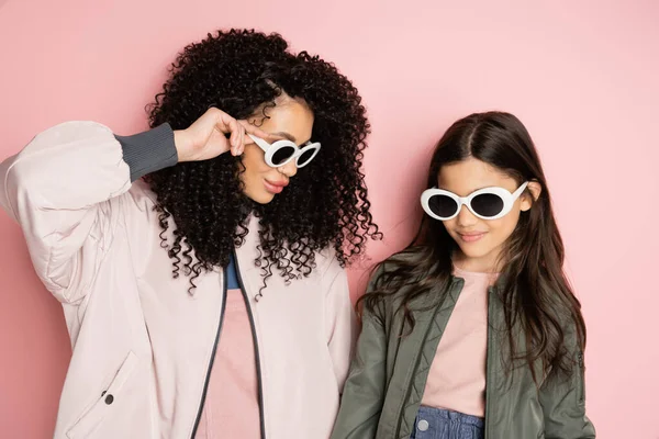 Donna alla moda che tiene gli occhiali da sole vicino a figlia su sfondo rosa — Foto stock