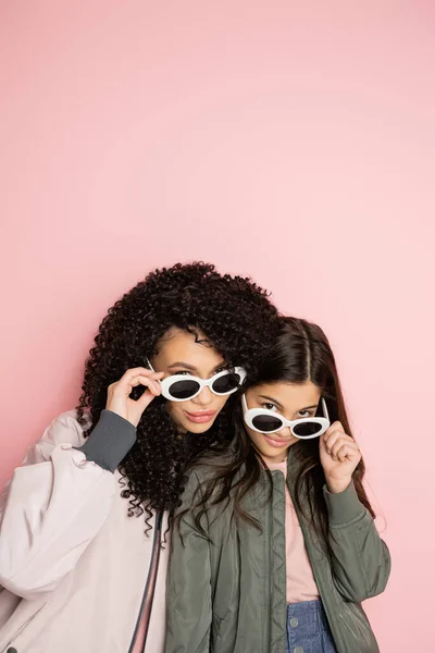 Elegante donna e bambino in occhiali da sole guardando la fotocamera su sfondo rosa — Foto stock