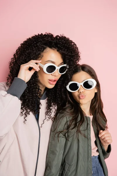 Preteen girl in sunglasses pouting lips near curly mom on pink background - foto de stock