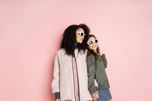 Cheerful preteen kid in sunglasses holding hand of stylish mom on pink background - foto de stock
